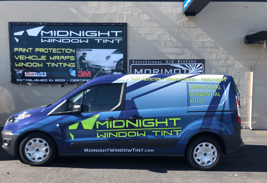 Side view of a customized van belonging to Midnight Window Tinting, parked in front of their shop, featuring vibrant graphics and services advertised such as window tinting, paint protection, and vehicle wraps.